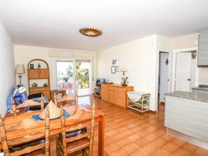 Een ruime woonkamer met een houten eettafel, een bank, een keuken met een wit aanrechtblad en een glazen schuifdeur naar een balkon.
