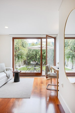 A modern living room with a large sliding glass door leading to a garden, a white sofa, a small black side table, and a wooden desk with a chair. - 1BR With Private Patio Porto Urban Flats (Porto)