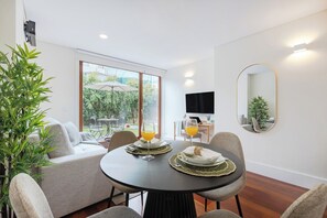 A dining area with a round table, chairs, and a sofa. There is a television on the wall and a large window with a view of greenery outside. - 1BR With Private Patio Porto Urban Flats (Porto)