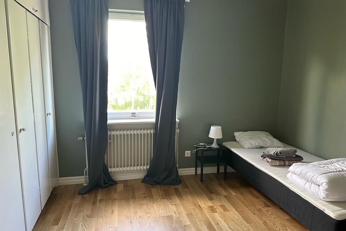 A bedroom with a single bed, a nightstand, a lamp, and a window with curtains.