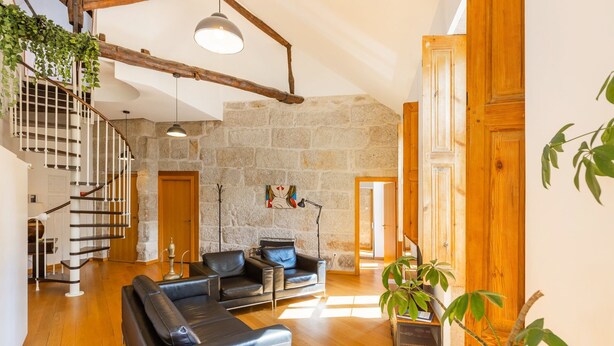 Amplia sala de estar con pared de piedra, vigas de madera, una escalera, sofás de cuero y una planta en maceta.