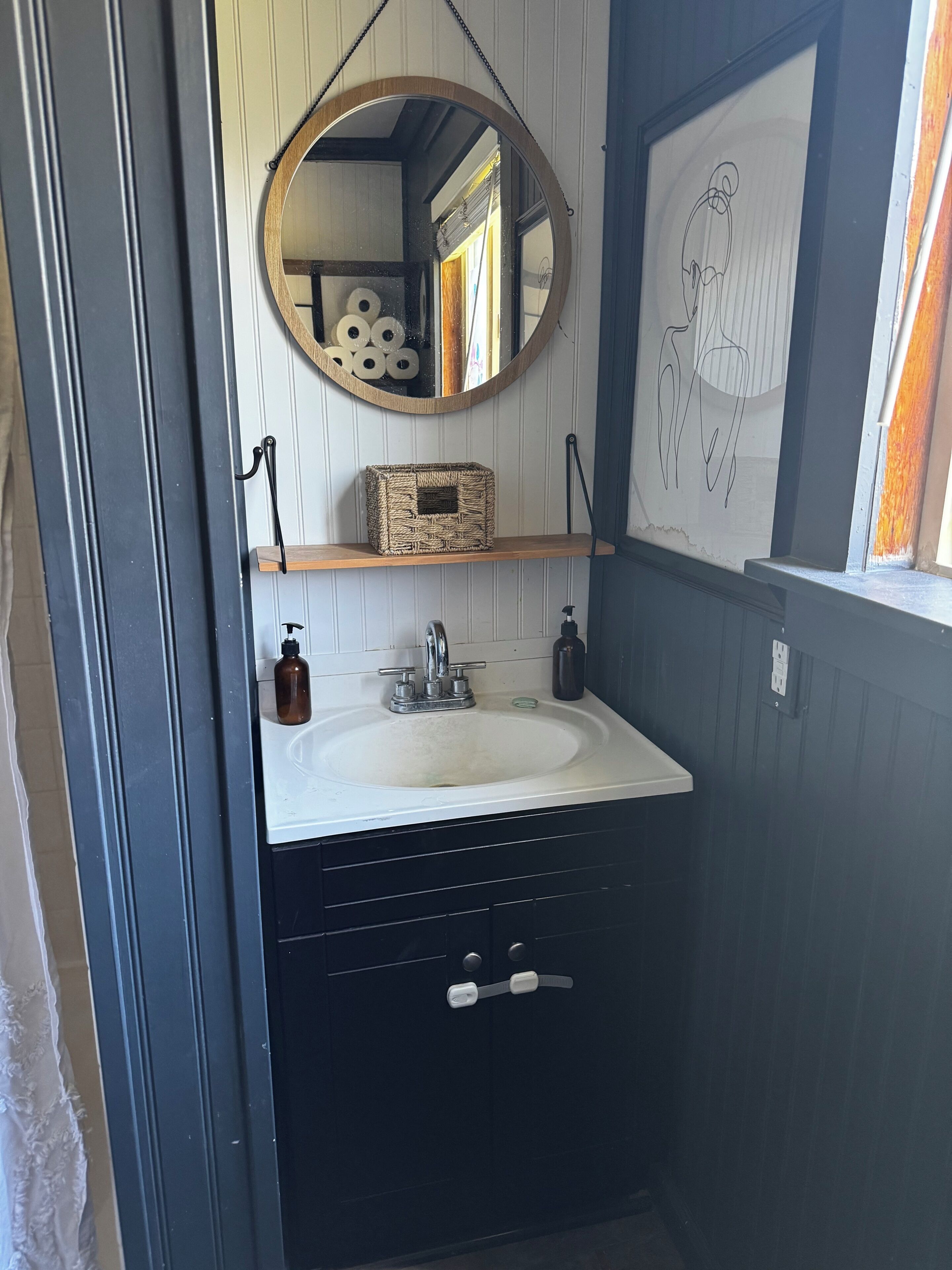 Une salle de bain avec un lavabo blanc, des meubles noirs, une étagère en bois sur laquelle repose un panier tressé, un miroir rond et une œuvre d’art encadrée accrochée au mur.