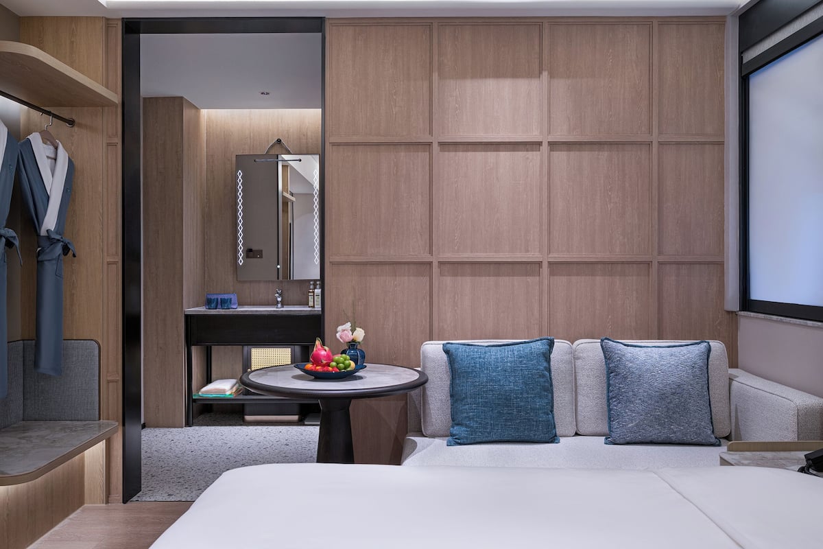 Une chambre d’hôtel moderne comprenant un lit, une table ronde avec un panier de fruits, un canapé avec des coussins bleus, un mur recouvert de panneaux en bois et une salle de bain avec un miroir et un porte-serviettes.
