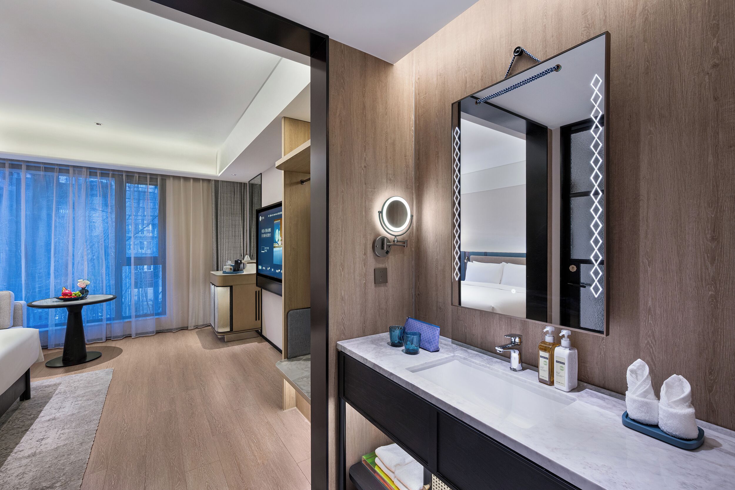 A modern hotel room with a large bed, a round mirror, a vanity with toiletries, and a view of the city through large windows.