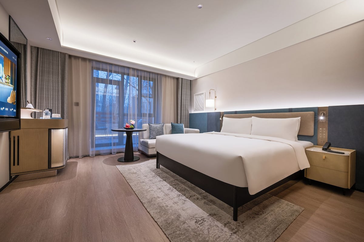 Une chambre d’hôtel moderne avec un grand lit, un bureau avec une télévision, une petite table avec une lampe et une vue sur l’extérieur.