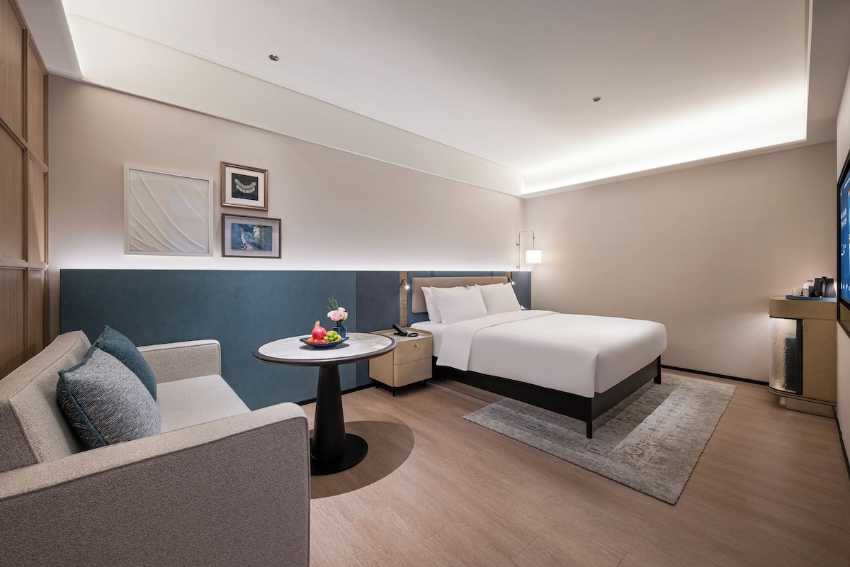 Une chambre d’hôtel moderne avec un lit, un canapé, une petite table ronde avec des fruits, une table de chevet et des œuvres d’art murales.