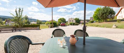 An outdoor seating area with a table and chairs under a large umbrella, overlooking a road with houses and greenery.