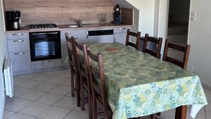 A dining area with a table covered in a floral tablecloth, surrounded by wooden chairs. A kitchen with modern appliances and a wooden countertop is visible in the background.