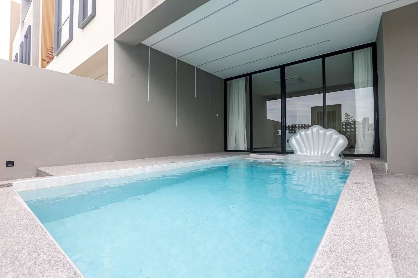 Ein Schwimmbecken mit klarem, blauem Wasser, einem weißen, muschelförmigen Schwimmdock und einem modernen Gebäude mit großen Fenstern sowie einer überdachten Terrasse.