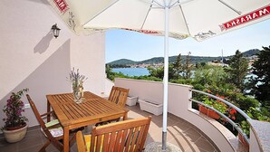 Ein Holztisch und Stühle unter einem weißen Sonnenschirm auf einem Balkon mit Blick auf das Meer und die Hügel.
