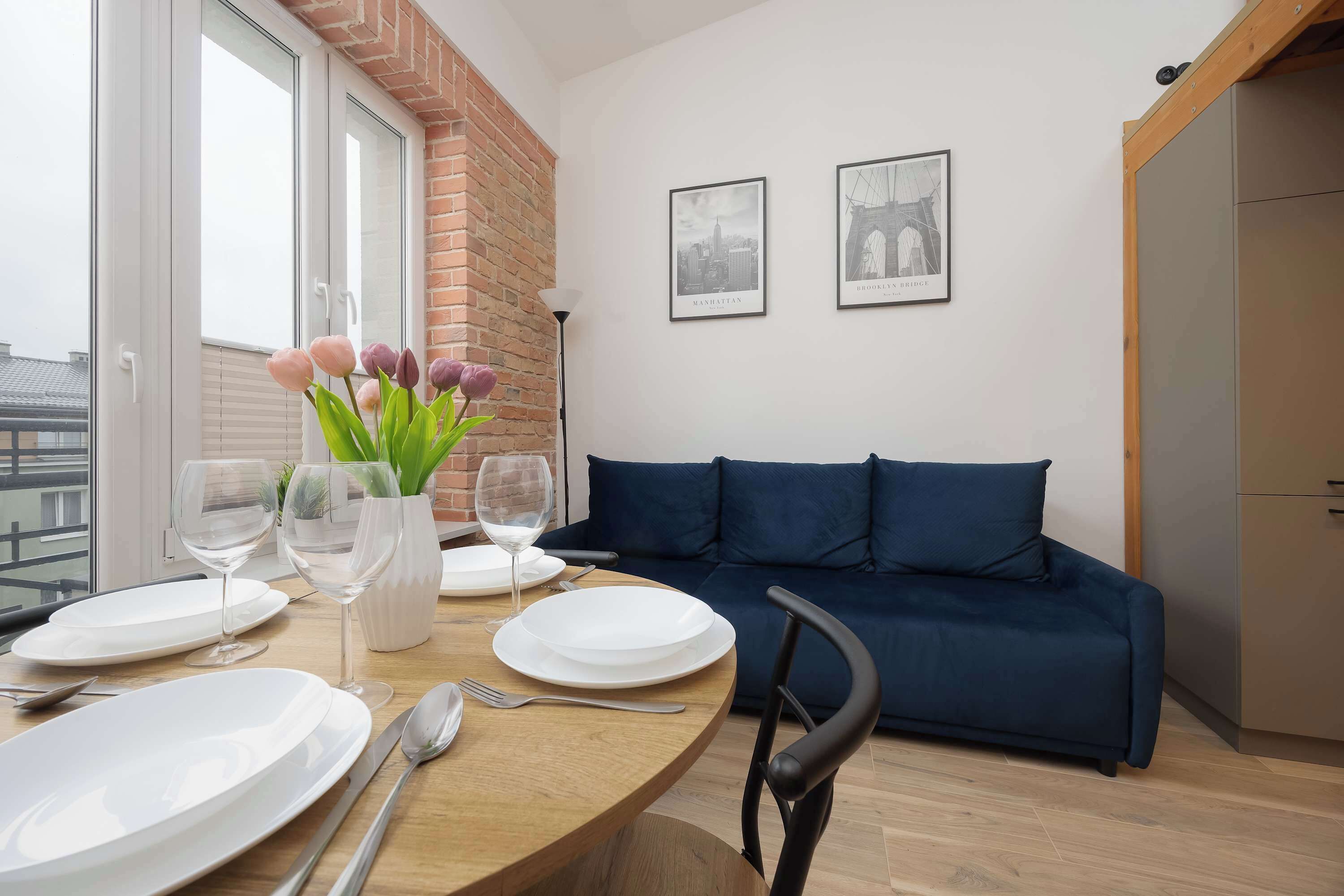Un coin repas avec une table ronde, de l’assiette blanche, des couverts et un vase rempli de fleurs. On y trouve également un canapé bleu et un tableau représentant le pont de Brooklyn, accroché au mur.