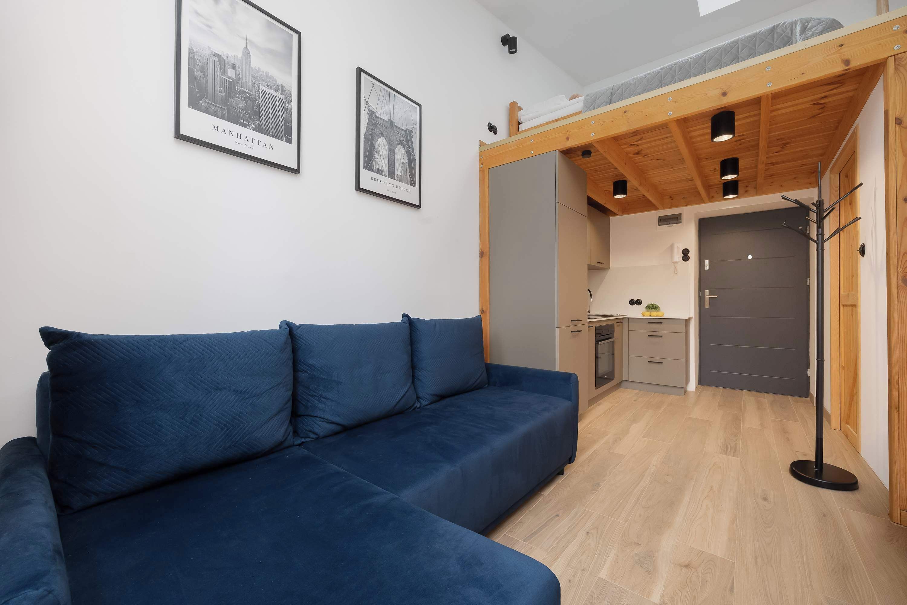 A modern room with a blue sofa, wooden ceiling beams, a kitchenette, and two framed pictures on the wall.