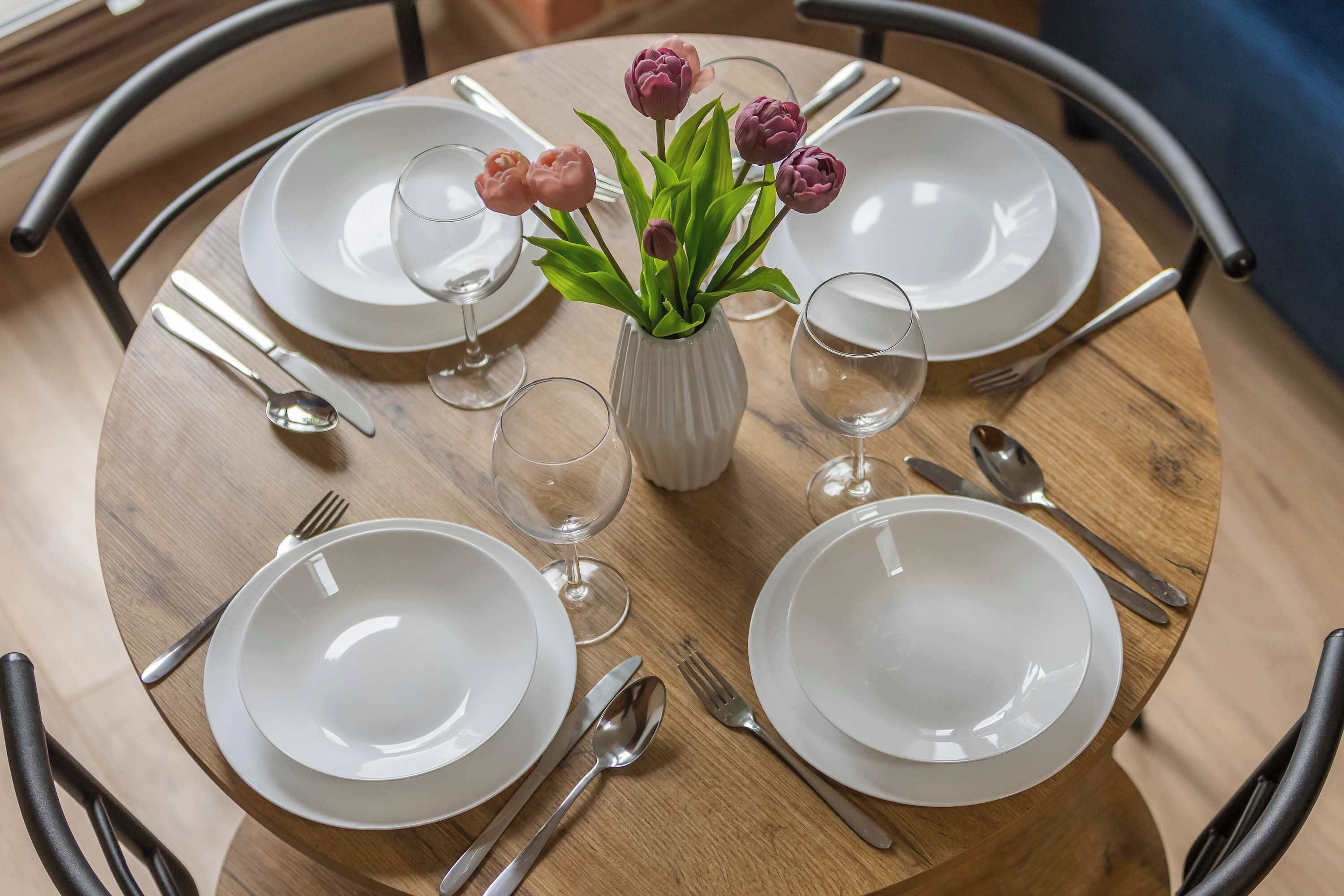 Mesa de madera redonda dispuesta para cuatro personas, con platos blancos, cubiertos y un florero con flores rosas.