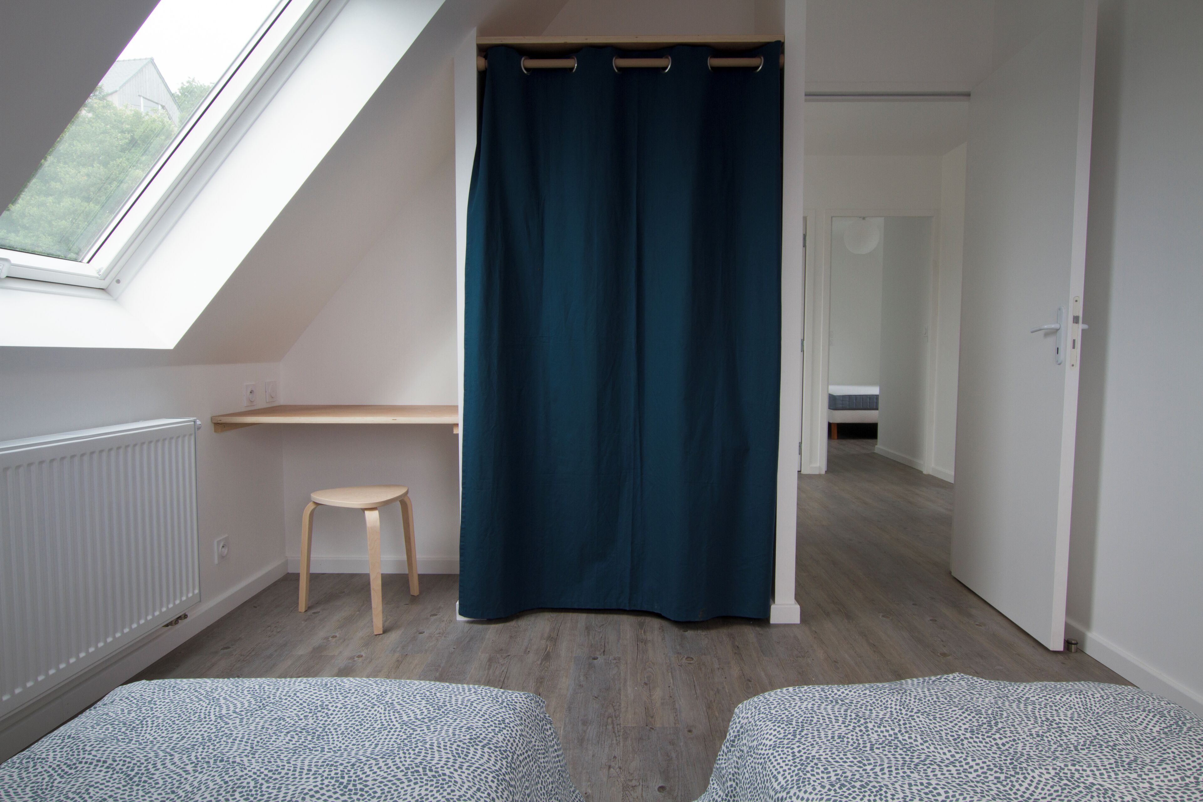 A room with a skylight, a dark curtain, a wooden stool, and a bed with patterned bedding.