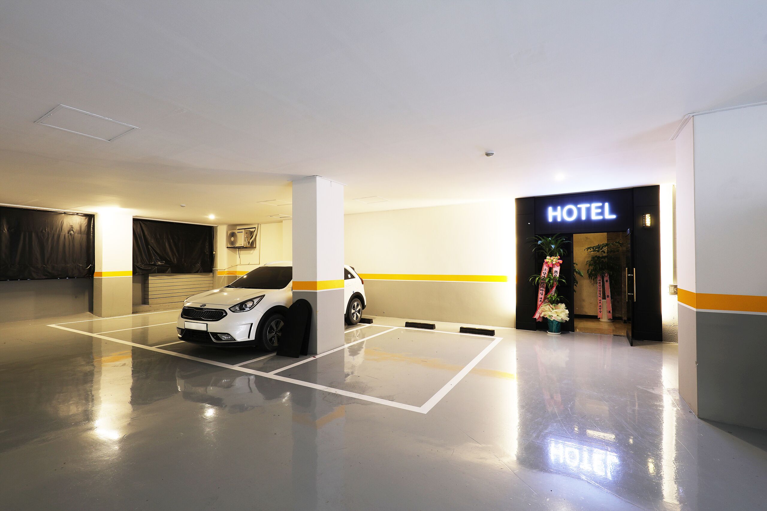A hotel parking area with a white car parked, a visible entrance, and a sign that reads 'HOTEL'.