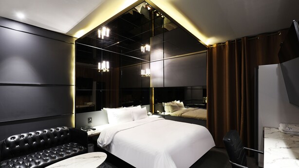 A modern hotel room with a large bed, a black leather sofa, a marble-topped table, and a large mirror. - Stay25 (Seongnam)