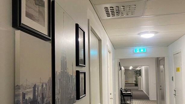 Un pasillo con cuadros de paisajes urbanos en la pared, una puerta y una mesa con sillas a lo lejos.