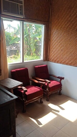 Una habitación con dos sillas de madera, una mesa, un ventanal con vistas a follaje y una pared de tejido.