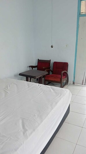 Una habitació amb un llit, una taula, dues cadires, una làmpada penjant i una porta.