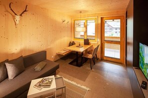 A cozy room with a wooden wall, a sofa, a dining table with chairs, a TV, and a window with a view of buildings. - Eichen Lodge Mellau (Mellau)