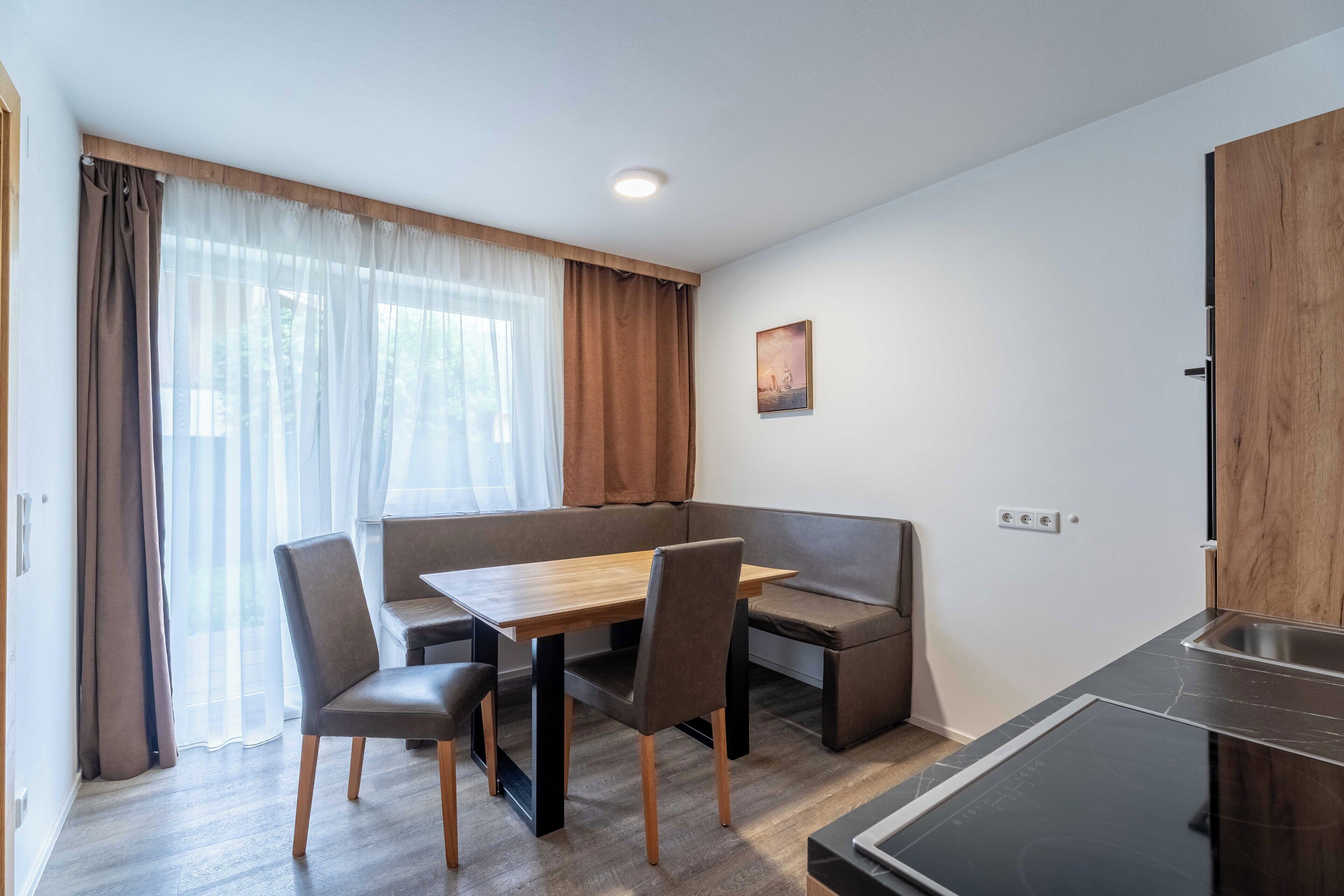 A modern room with a dining table, chairs, a sofa, a small kitchenette, and a window with curtains.