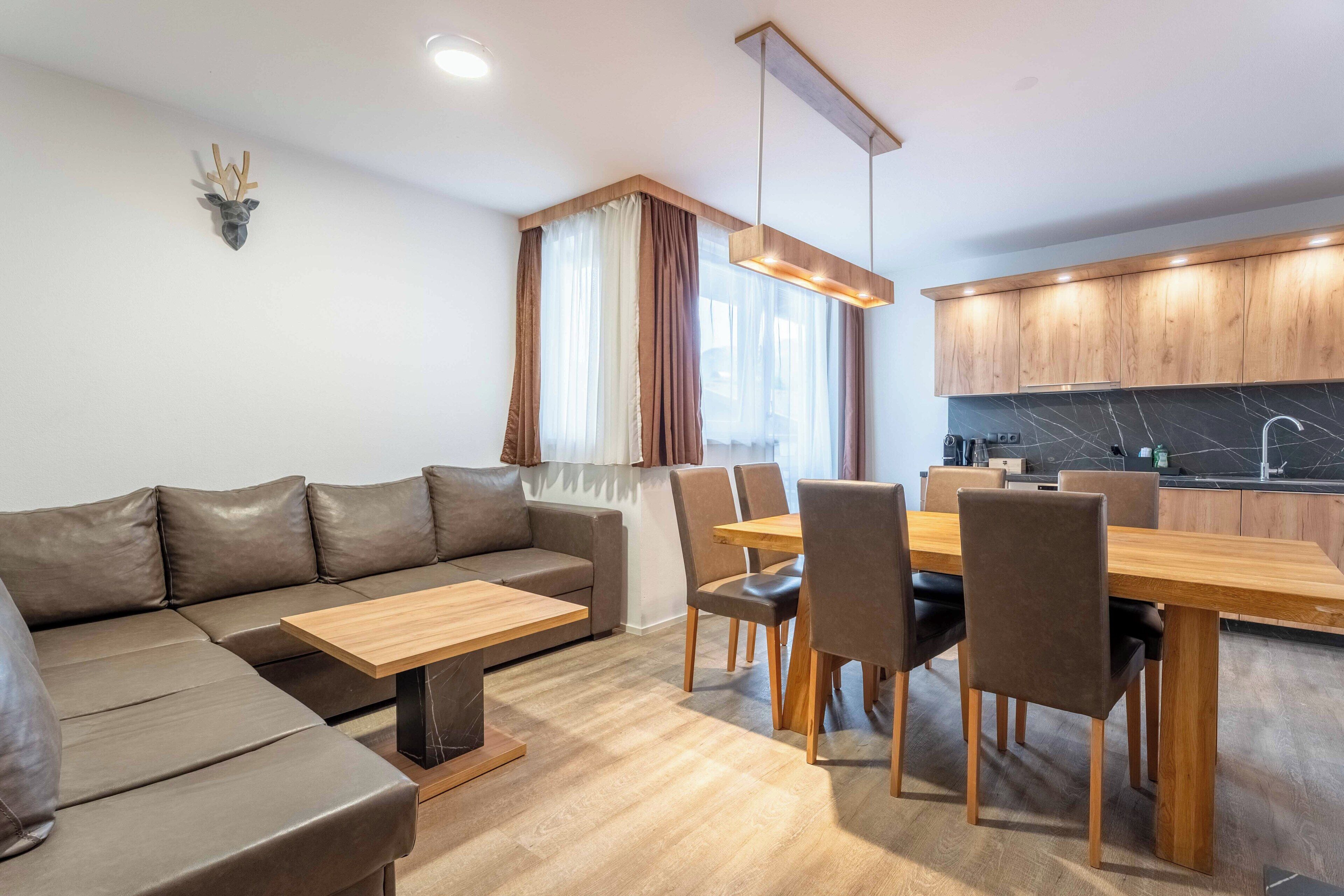 A modern living room with a sofa, dining table, chairs, and a kitchen area with wooden cabinets and a marble backsplash.