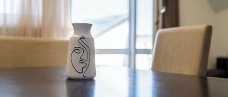 Eine weiße Vase mit einer schwarzen Linienzeichnung auf einem Tisch in einem Zimmer mit Fenster und Stuhl.