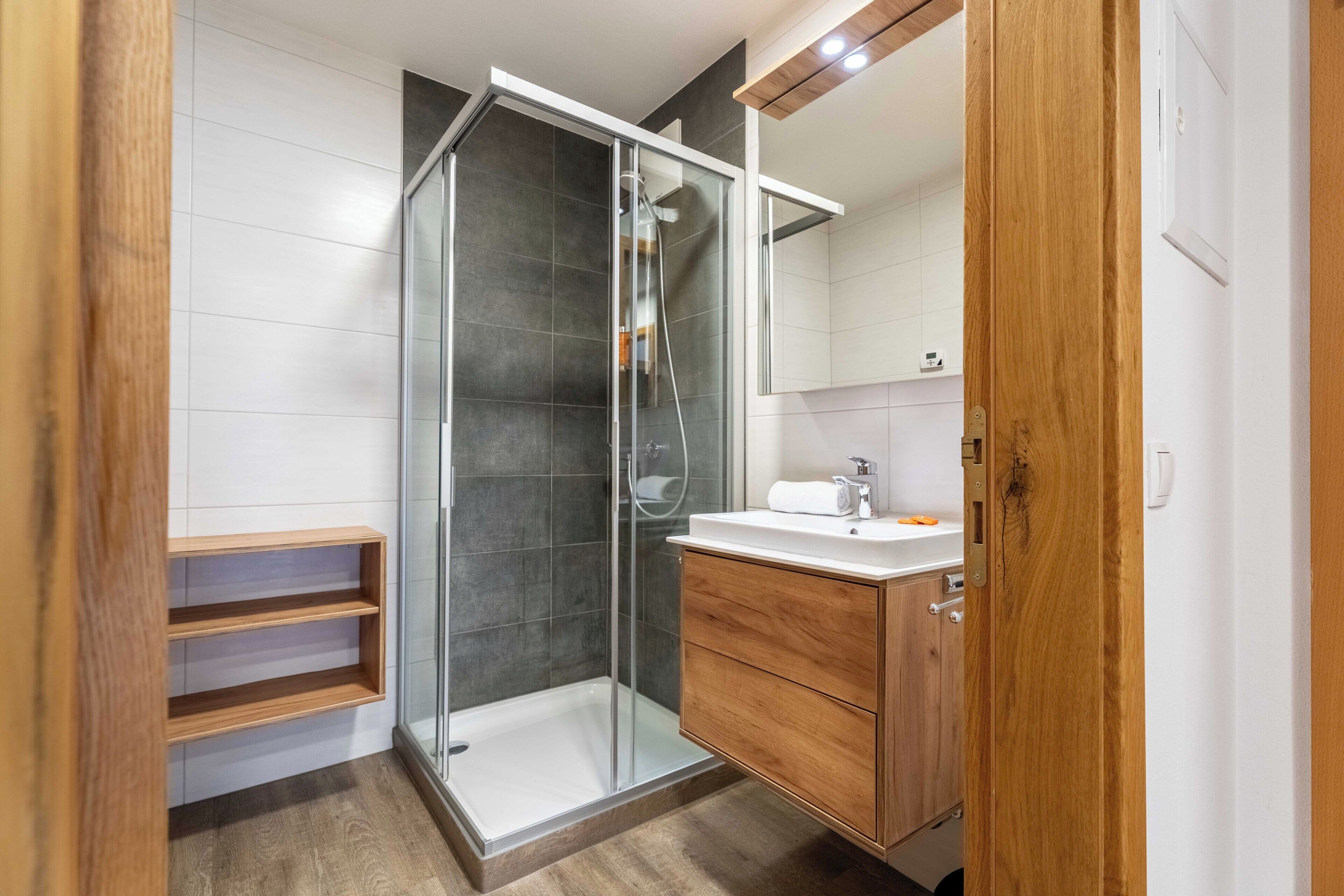 A modern bathroom with a glass shower enclosure, wooden shelves, a white sink, and a wooden vanity.