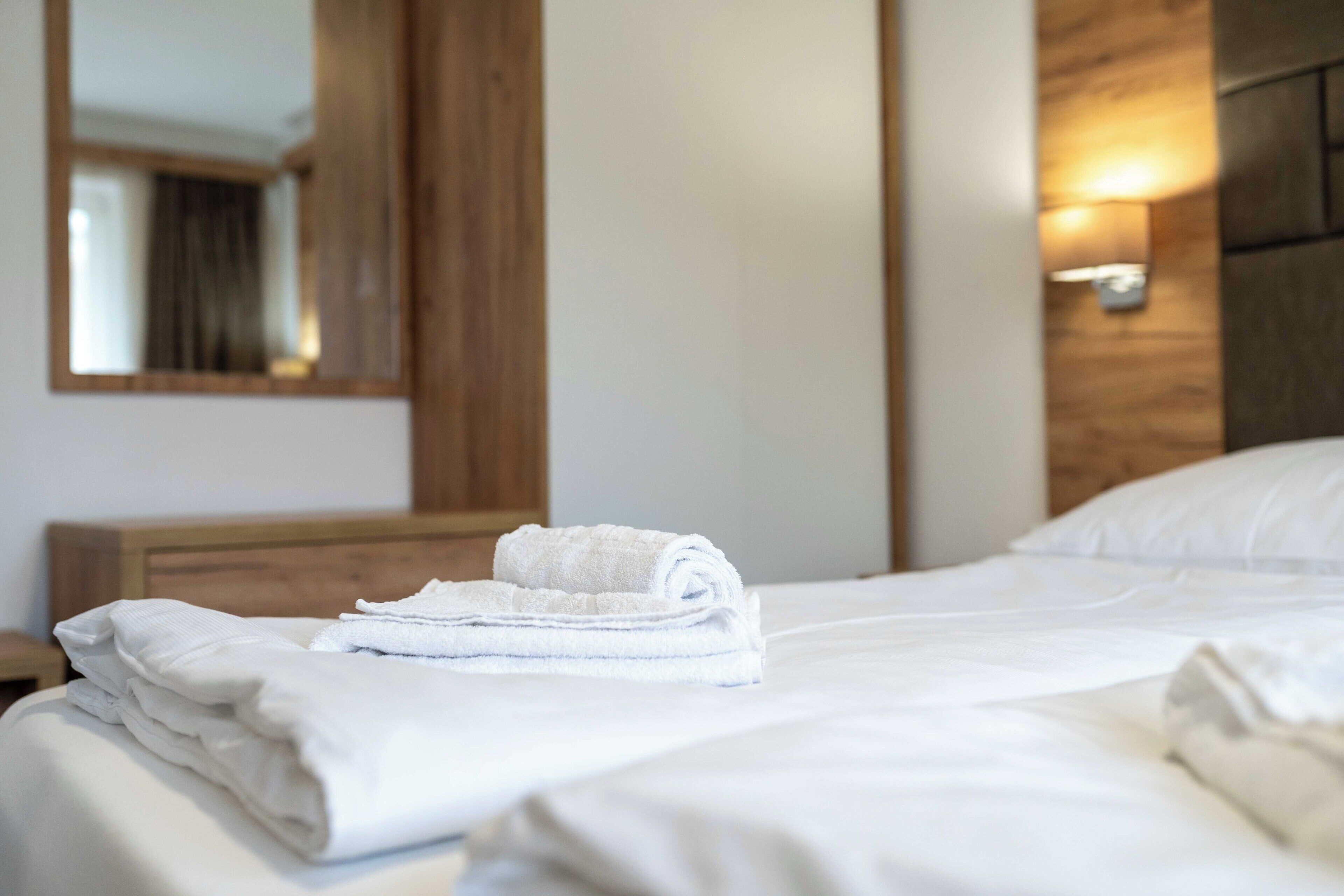 Una cama bien hecha con toallas blancas dobladas, cabecera de madera, una lámpara de pared y un espejo que refleja un rincón de la habitación.