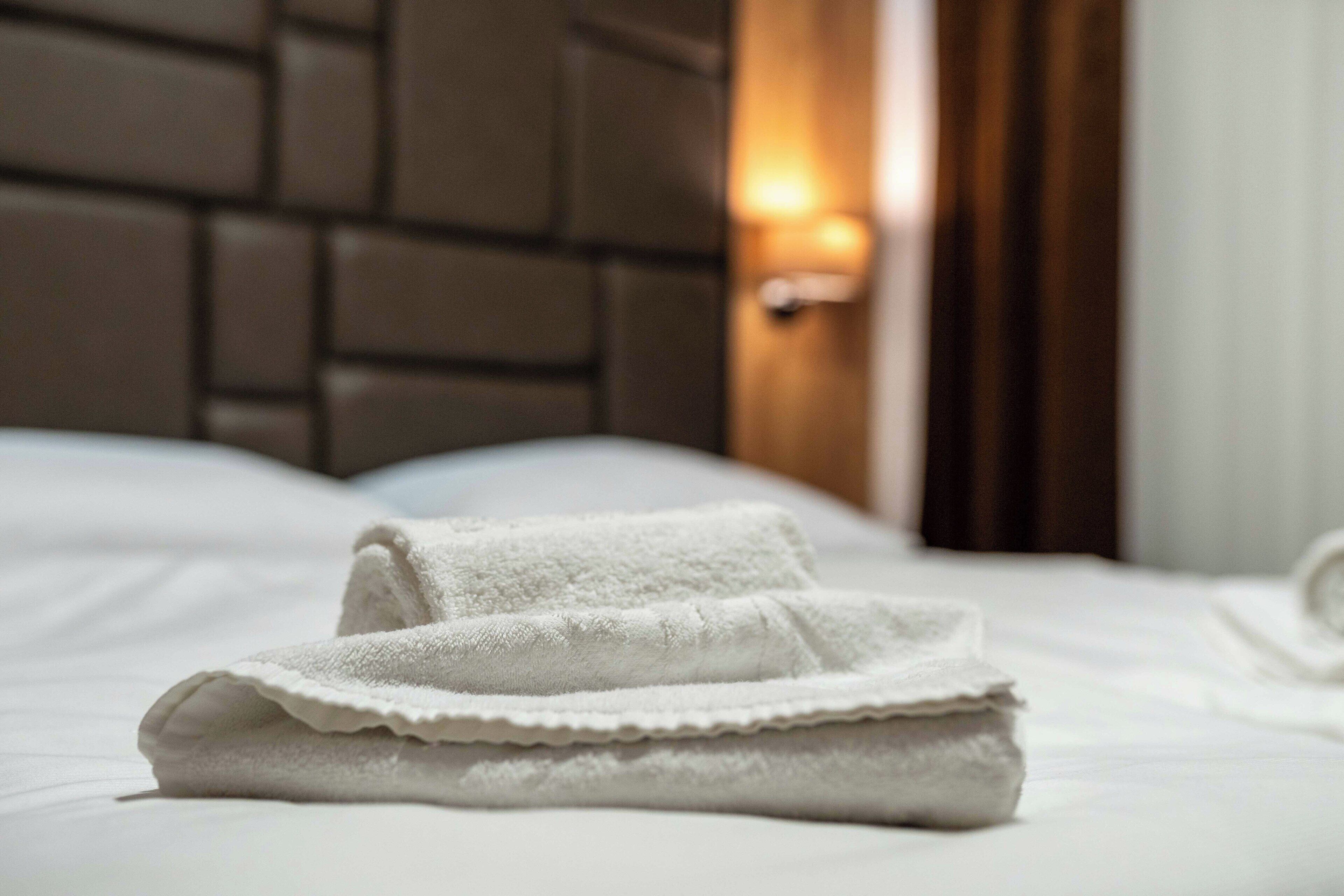 A neatly folded white towel on a bed with a headboard and a wall-mounted light fixture.