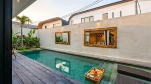 A swimming pool with a wooden deck, a fruit basket, and a modern building with large windows. - K Club Cemagi Villa 5 (Badung)