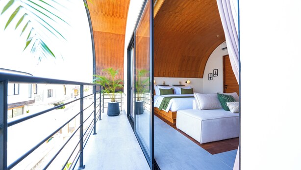 Ein Balkon mit Metallgeländer, ein Bett mit weißer Bettwäsche und grünen Kissen sowie ein Blumentopf.