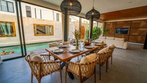 Ein Essbereich mit Blick auf einen Pool, mit Korbstühlen, einem Holztisch für sechs Personen und einem modernen Wohnbereich mit Fernseher und Wandverkleidung mit Muster.