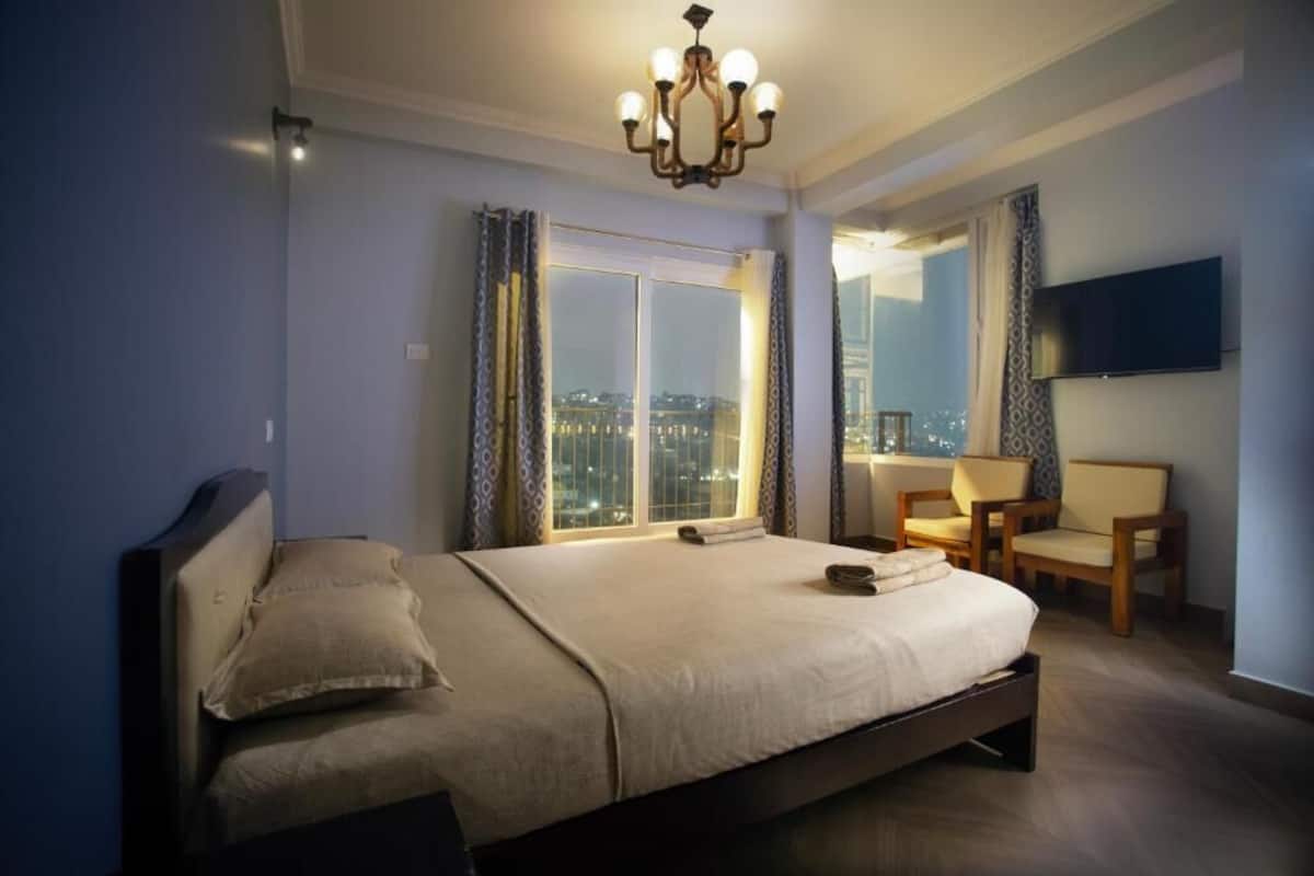 A bedroom with a large bed, a chandelier, a TV, and a city view.