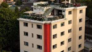 Un hotel a più piani con una facciata rossa e bianca ben visibile, una terrazza sul tetto con posti a sedere all'aperto e vista su un paesaggio collinare sullo sfondo.