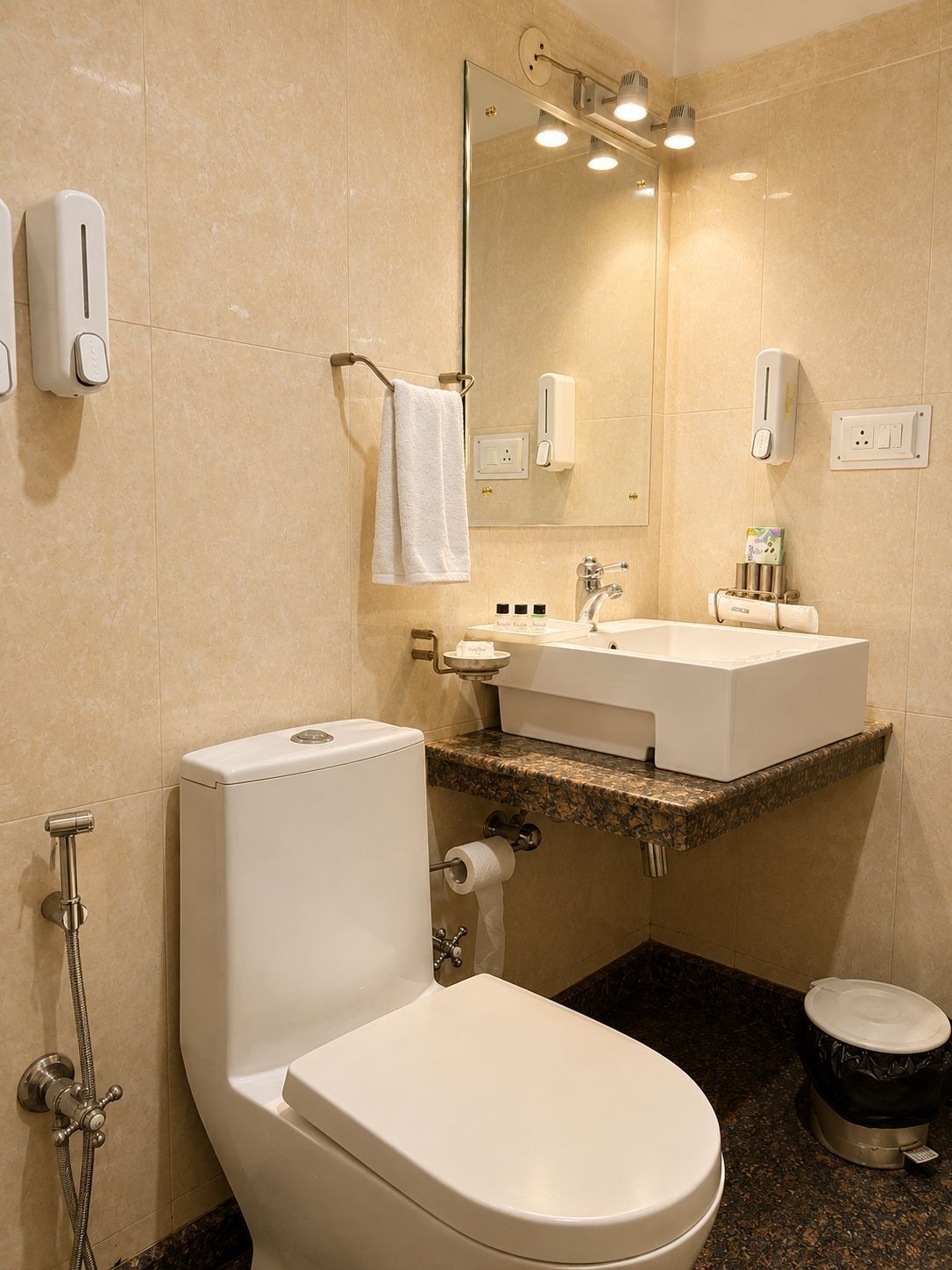 A bathroom with a toilet, bidet, sink, mirror, towel rack, and soap dispensers.
