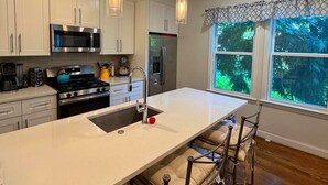 A modern kitchen with white cabinets, stainless steel appliances, a large island with bar stools, and a view of greenery through the window.