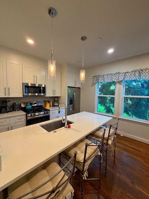 Una cocina moderna con armarios blancos, electrodomésticos de acero inoxidable, una isla grande con taburetes y vistas a la vegetación a través del ventanal.