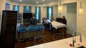A modern kitchen with a central island, a dining area with blue chairs, and a TV in the background.