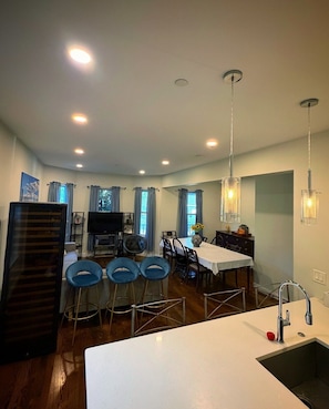 Una cocina moderna con una isla central, un comedor con sillas azules y un televisor al fondo.
