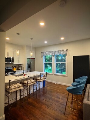 A modern kitchen with a central island, bar stools, and a dining area with a view of greenery.