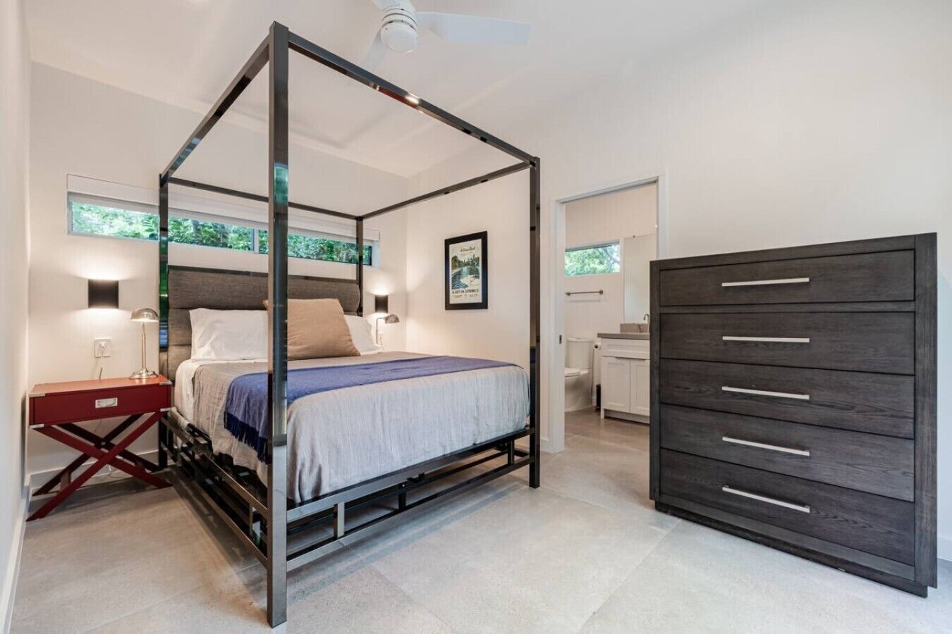 A modern bedroom with a four-poster bed, a bedside table with a lamp, a dresser with drawers, and a view of the outdoors through a window.