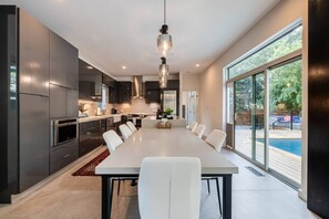 Una cocina moderna con una larga mesa de comedor, electrodomésticos integrados y un amplio ventanal con vistas a una piscina.