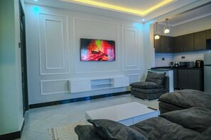 Een moderne woonkamer met een flatscreen televisie, een donkere bank en een keuken met donkere kasten.