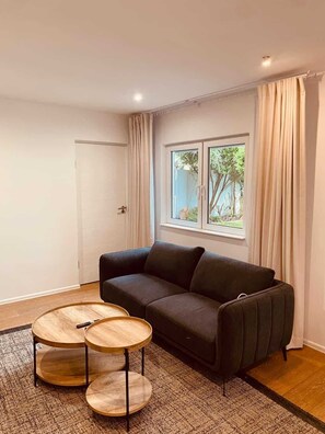 Ein modernes Wohnzimmer mit einem dunklen Sofa, zwei runden Holztischen, einem großen Fenster mit Vorhängen und einer Tür.