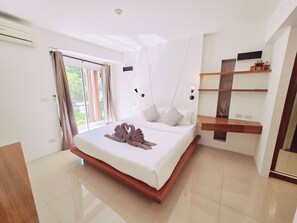 A modern bedroom with a large bed, a built-in shelf, and a view of greenery outside. - Replay Comfortable Apartment (Bophut)