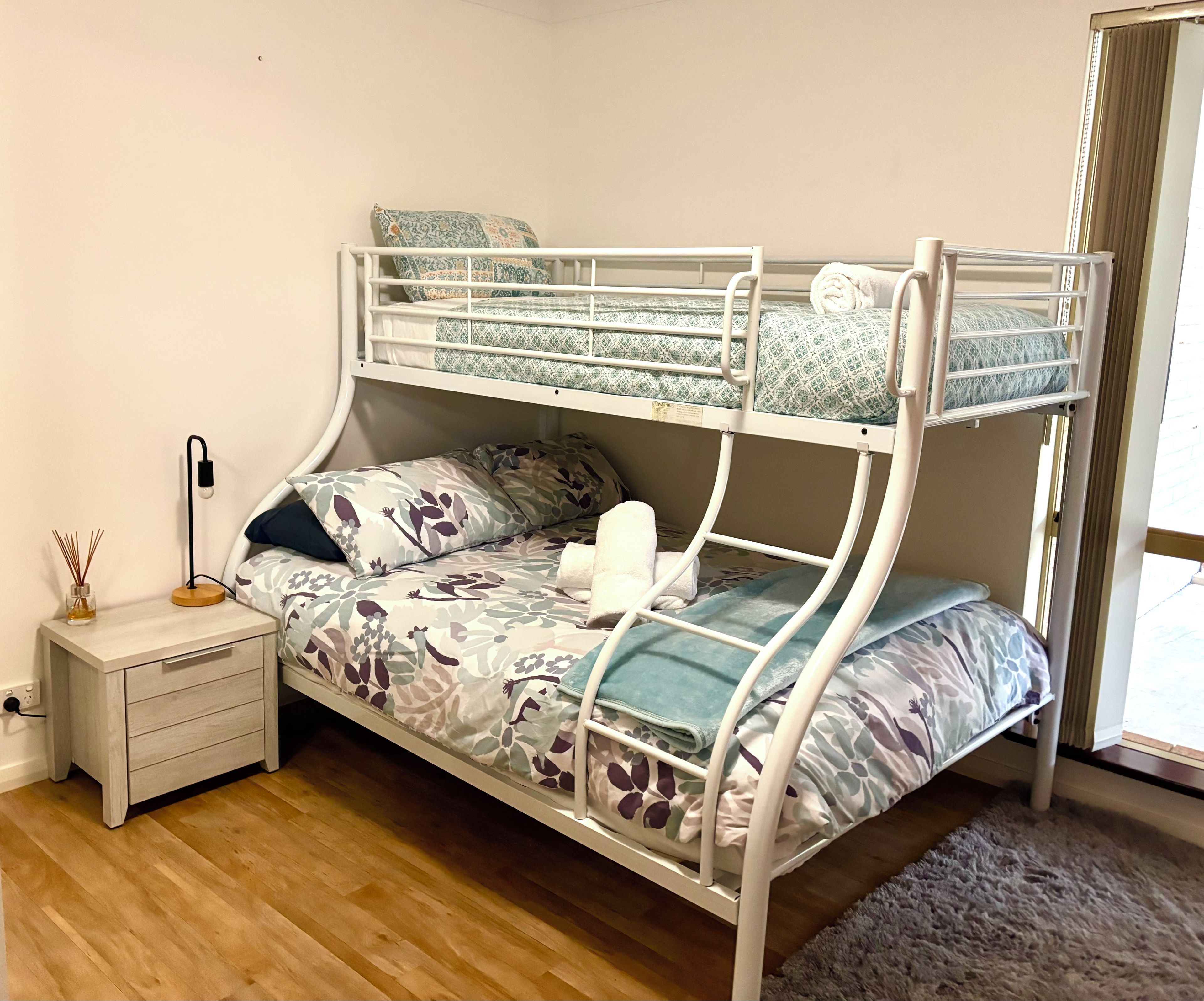 A bunk bed with floral bedding, a bedside table with a lamp, and a window with curtains.