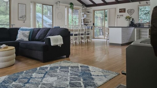 A spacious living area with a dark-colored sofa, a patterned rug, wooden flooring, and a kitchen area with white cabinets.