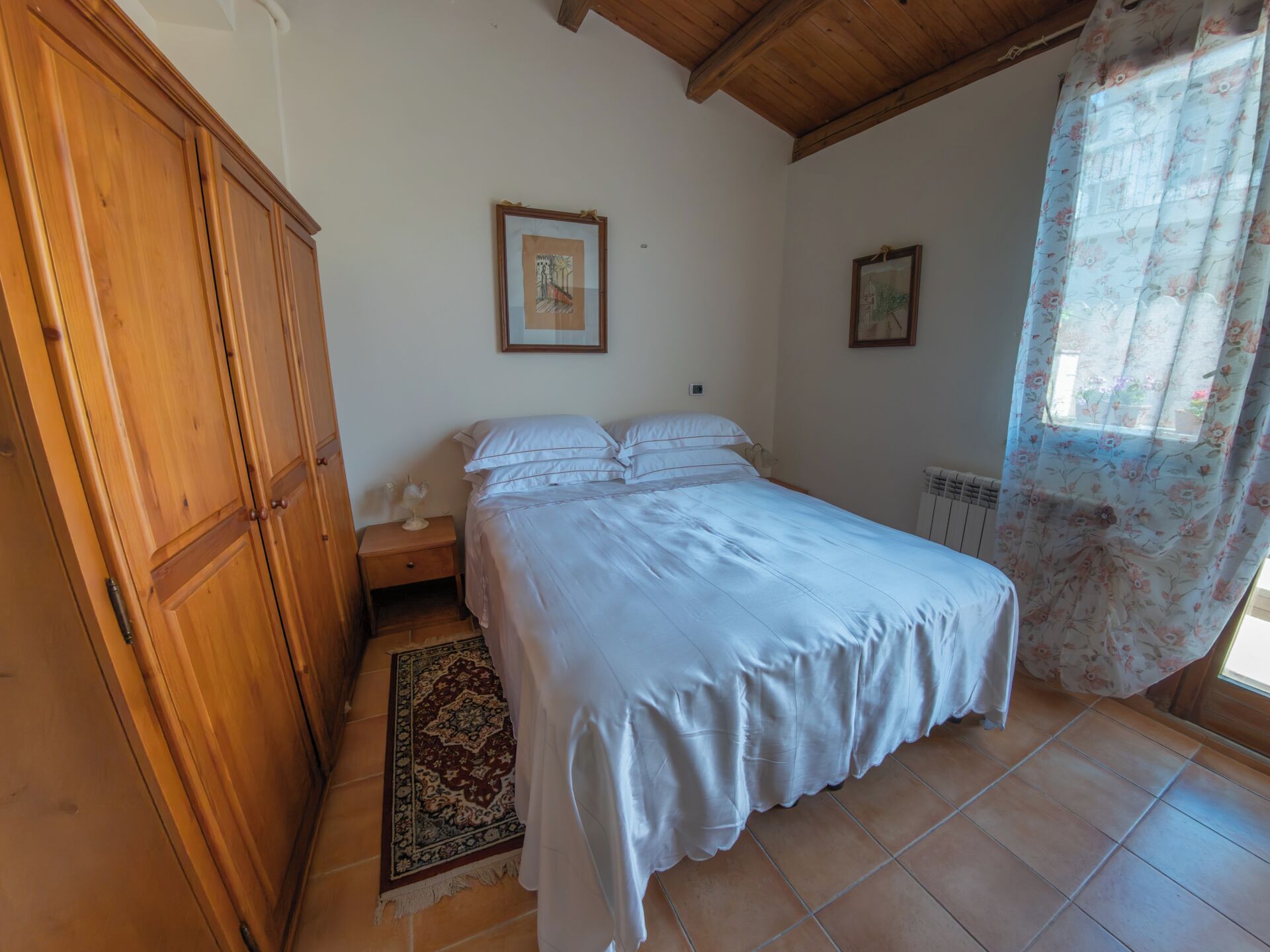 Une chambre à coucher avec un lit, une armoire en bois, deux tableaux encadrés, une table de chevet et une fenêtre ornée de rideaux à motifs floraux.