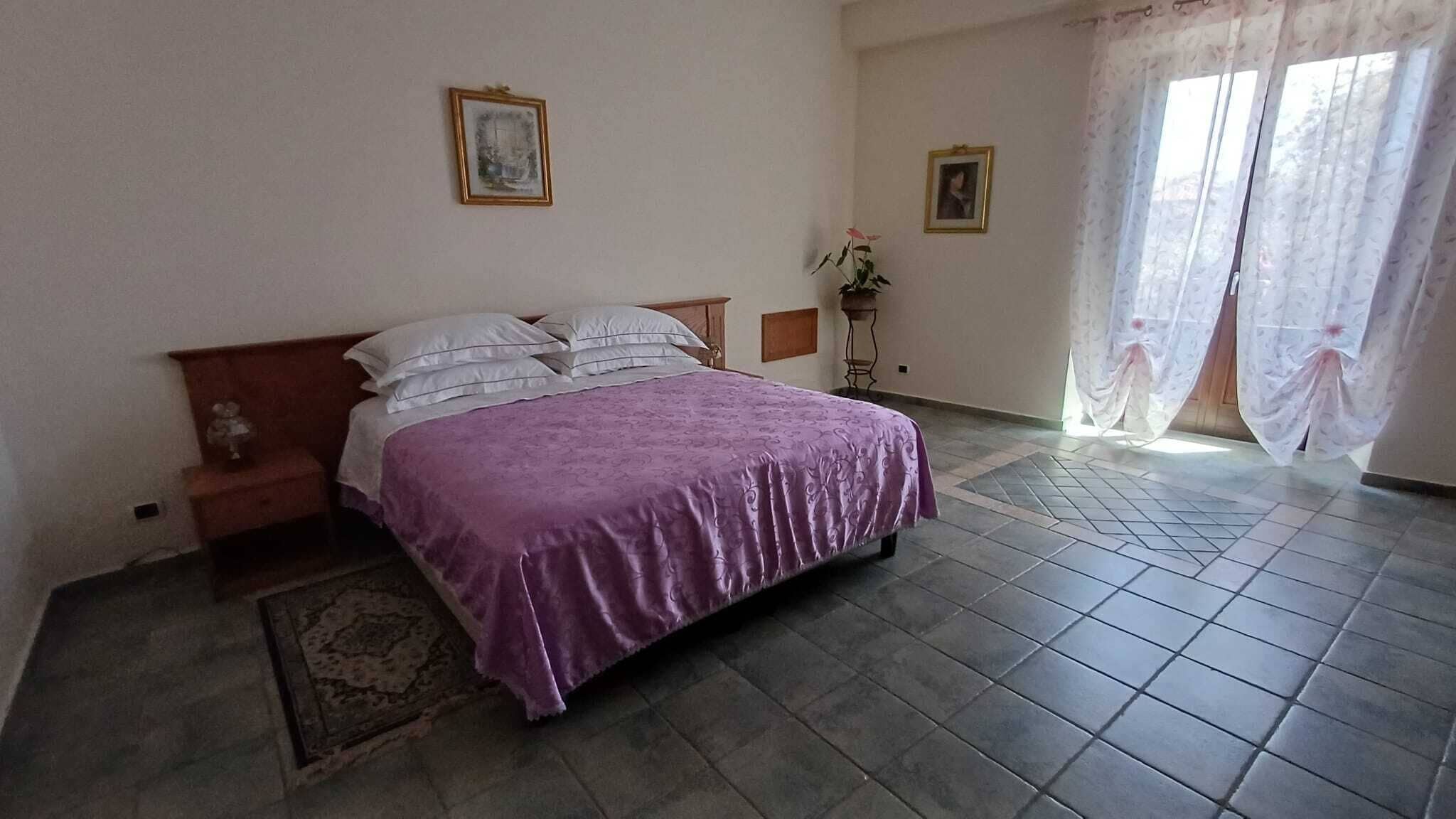 Um quarto com cama, mesa de cabeceira, mesinha, cadeira, tapete e janela com cortinas.